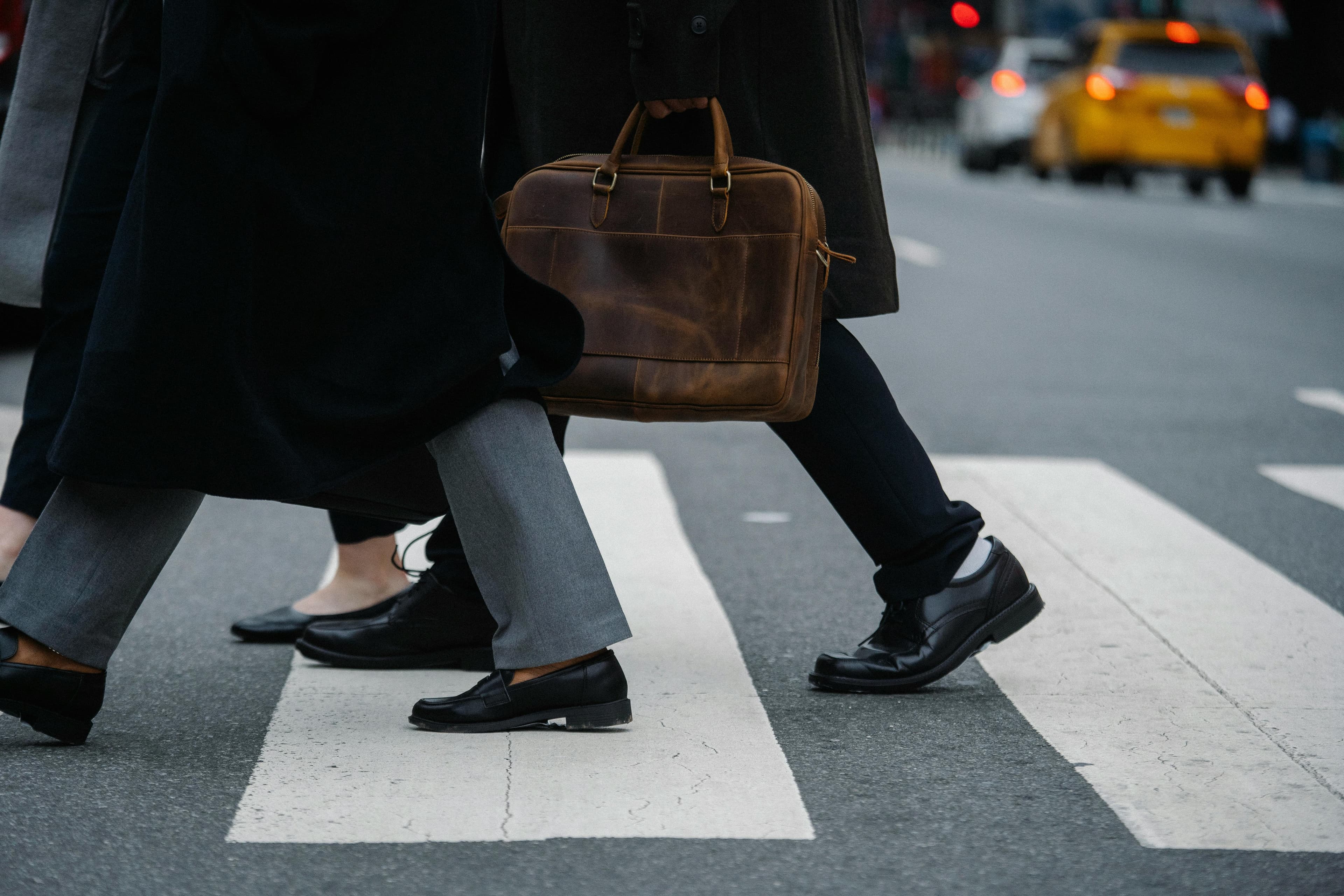 Professionals crossing a city street, briefcase in focus, taxi in background