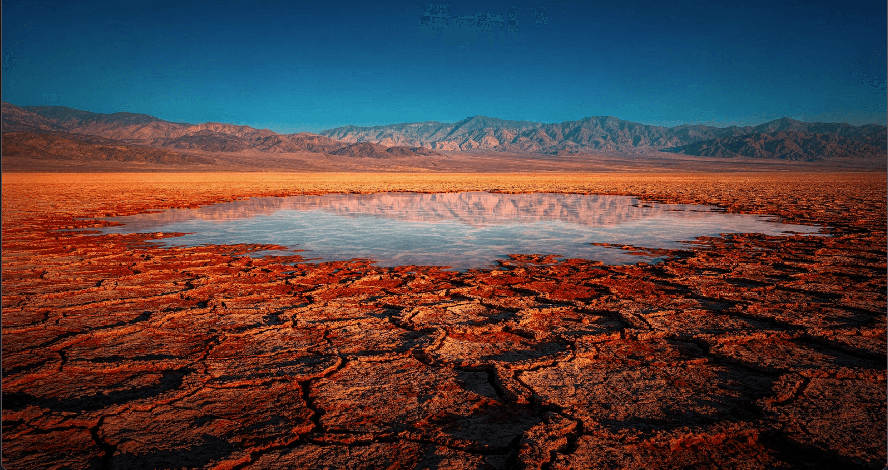 Cracked desert salt flat with a reflecting pool and mountains at dusk